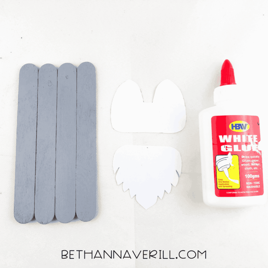 Craft stick base painted gray next to white paper pieces and glue