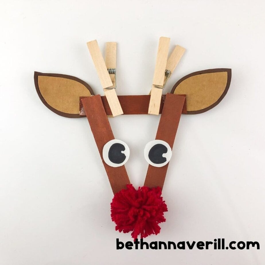 Completed reindeer face craft made from craft sticks, googly eyes, clothespins as antlers, paper ears, and a red yarn pom-pom nose