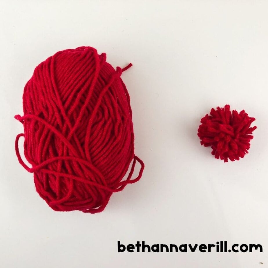 A skein of red yarn sits beside a small red pom-pom made from the same yarn.