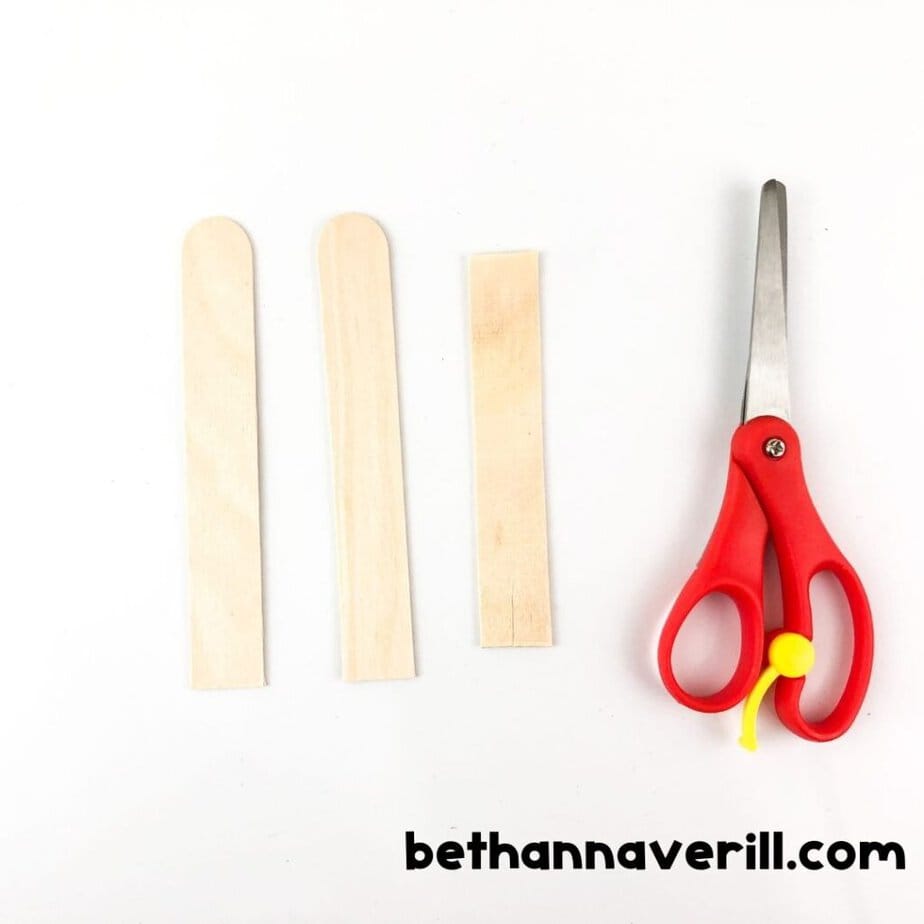 Two wooden craft sticks and one shorter stick lie next to red-handled scissors, ready for crafting.