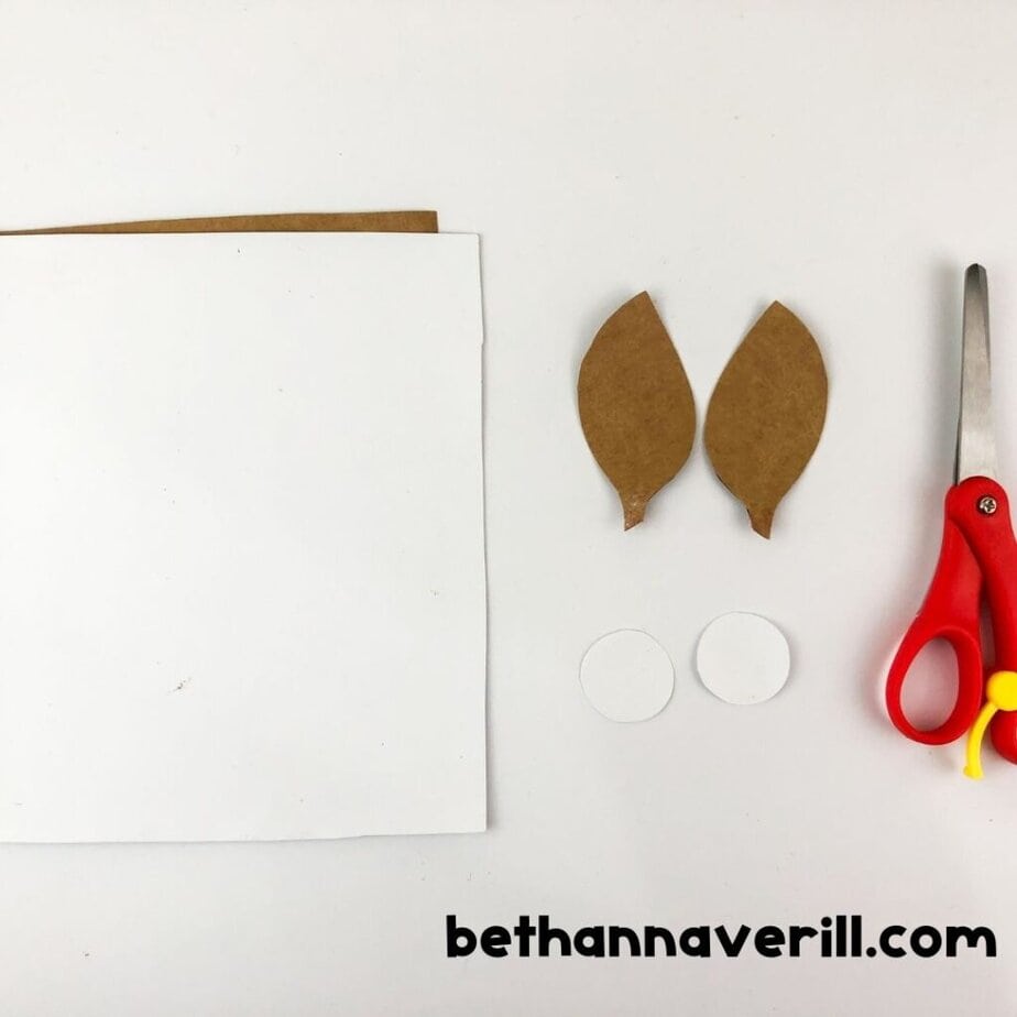 White paper, two tan ear shapes, two white eye circles, and scissors are laid out as crafting materials.
