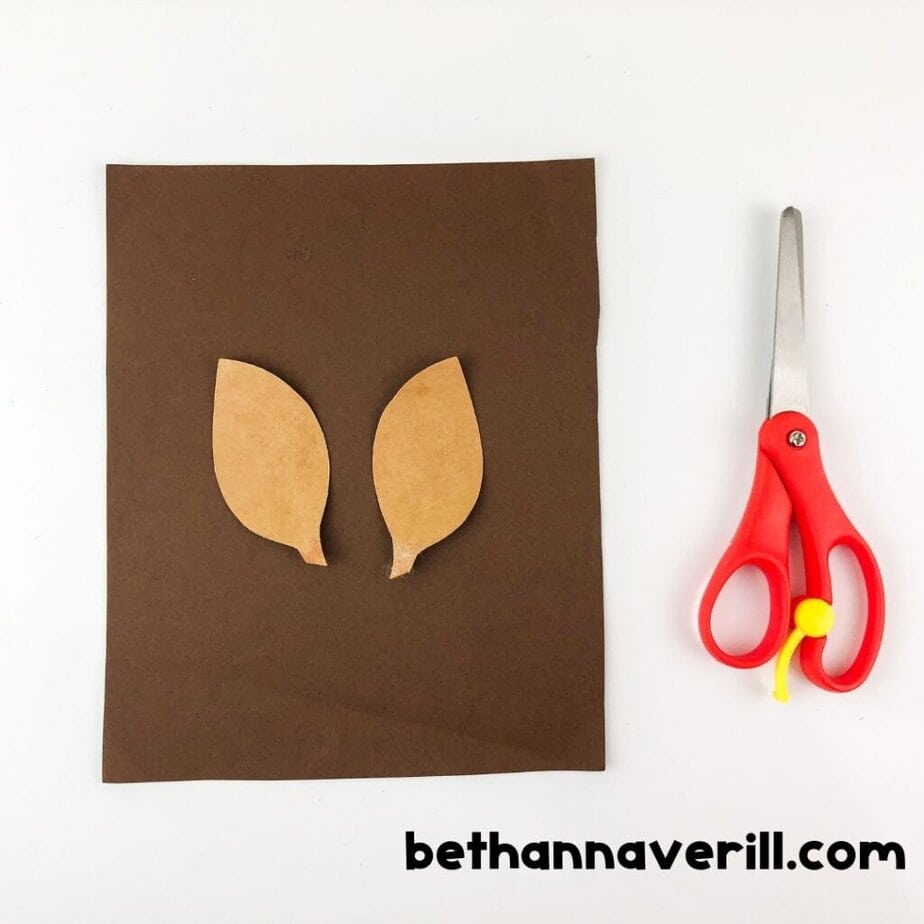 Two tan paper ear shapes are cut out and placed on a sheet of brown paper, next to a pair of red-handled scissors.