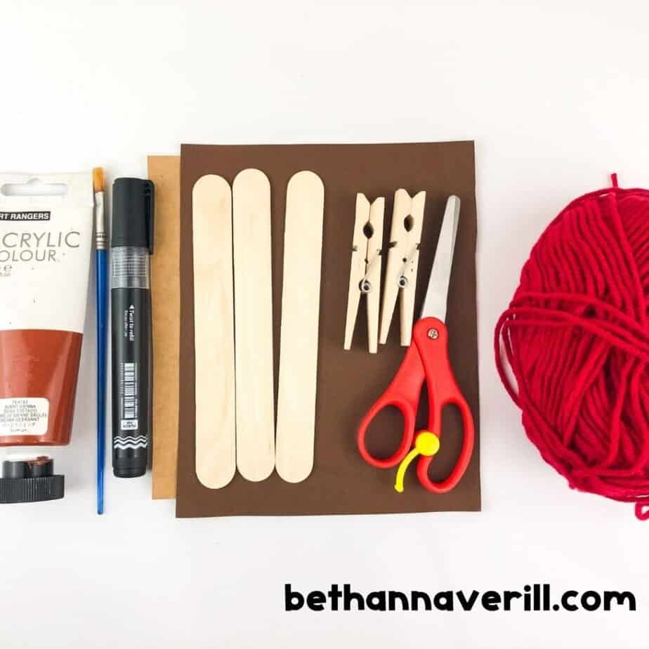 Craft supplies laid out, including brown and tan paper, red yarn, clothespins, craft sticks, paint, a black marker, scissors, and a paintbrush.