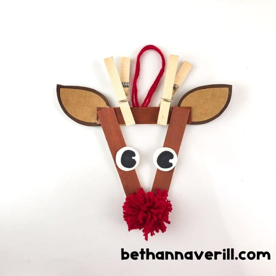 Completed reindeer ornament made from popsicle sticks, paper ears, googly eyes, clothespin antlers, a red yarn pom-pom nose, and a red yarn loop for hanging.