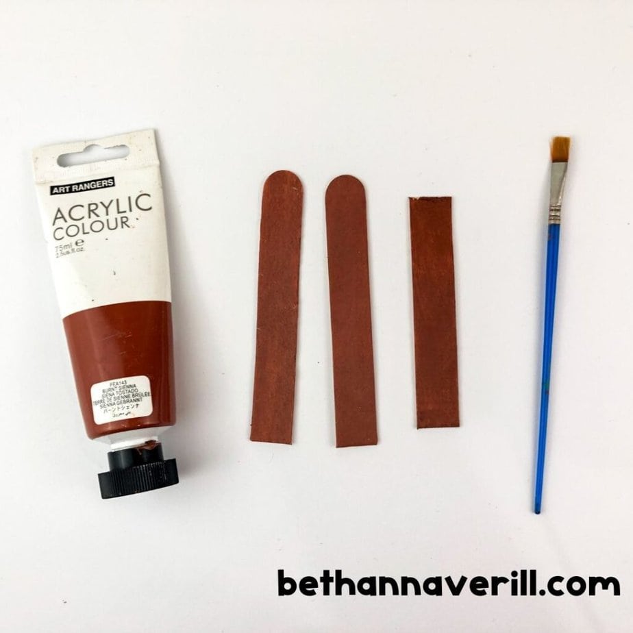 Two craft sticks and one short stick painted brown are laid beside a tube of brown acrylic paint and a flat brush.