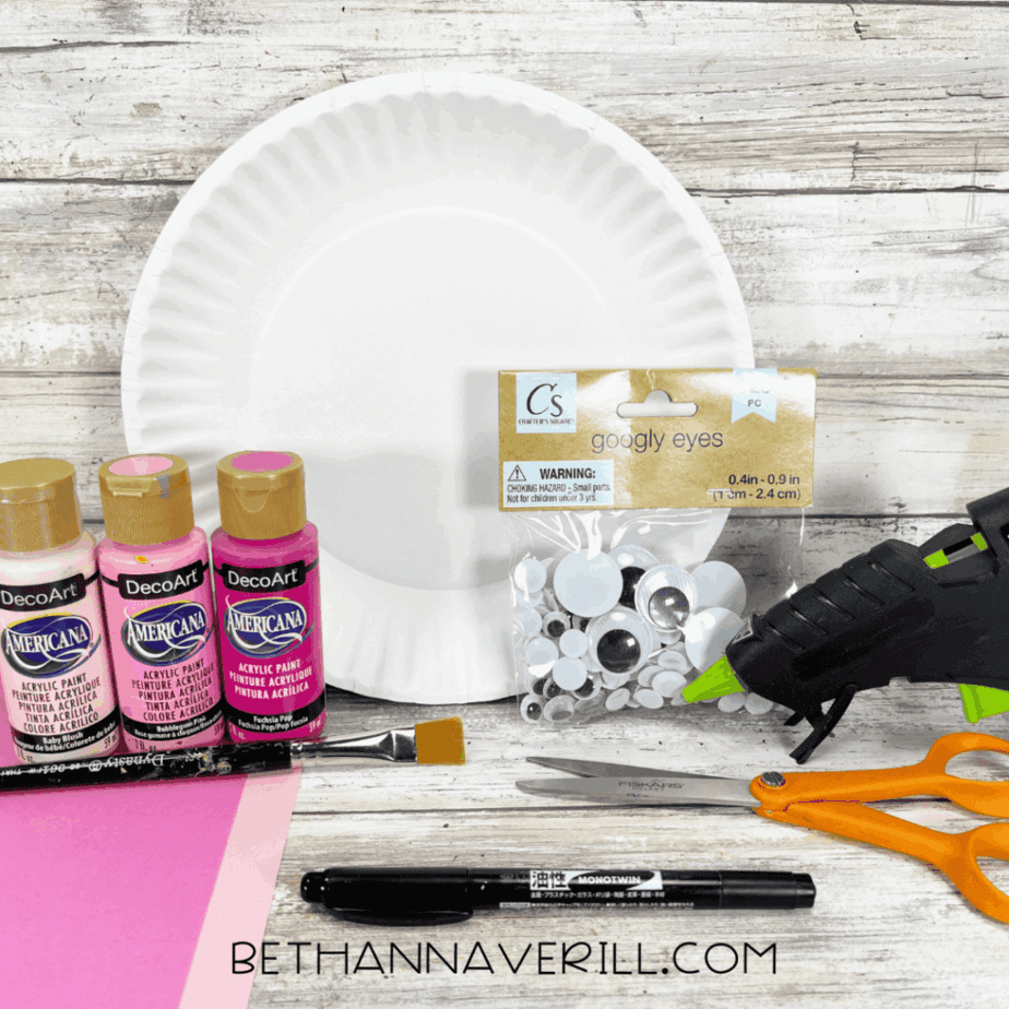 Materials for axolotl paper plate craft including paint, googly eyes, scissors, glue gun, and construction paper.