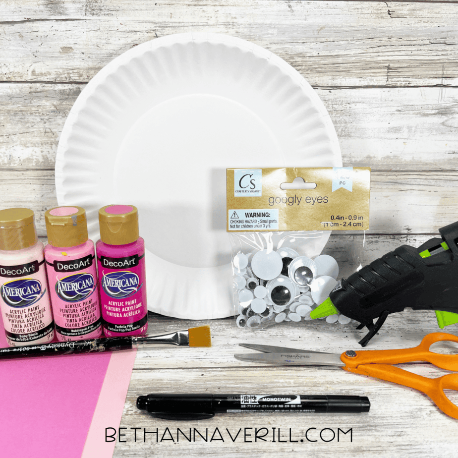 Materials for axolotl paper plate craft including paint, googly eyes, scissors, glue gun, and construction paper.