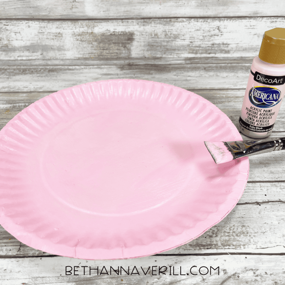 Paper plate painted pink with paintbrush and pink paint bottle.