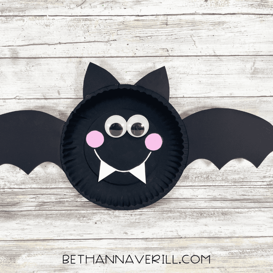 Finished bat paper plate craft with black wings, pointy ears, googly eyes, pink cheeks, and white fangs.
