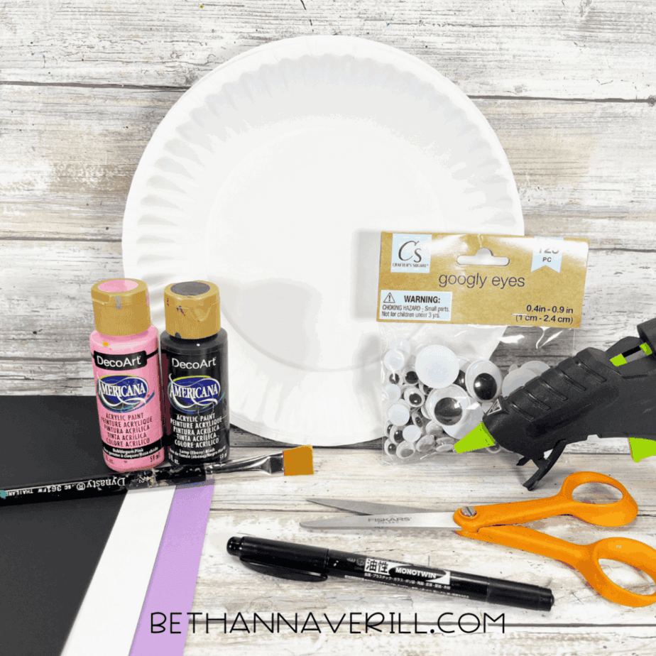 Materials for bat paper plate craft including paper plate, paints, googly eyes, scissors, glue gun, and markers.