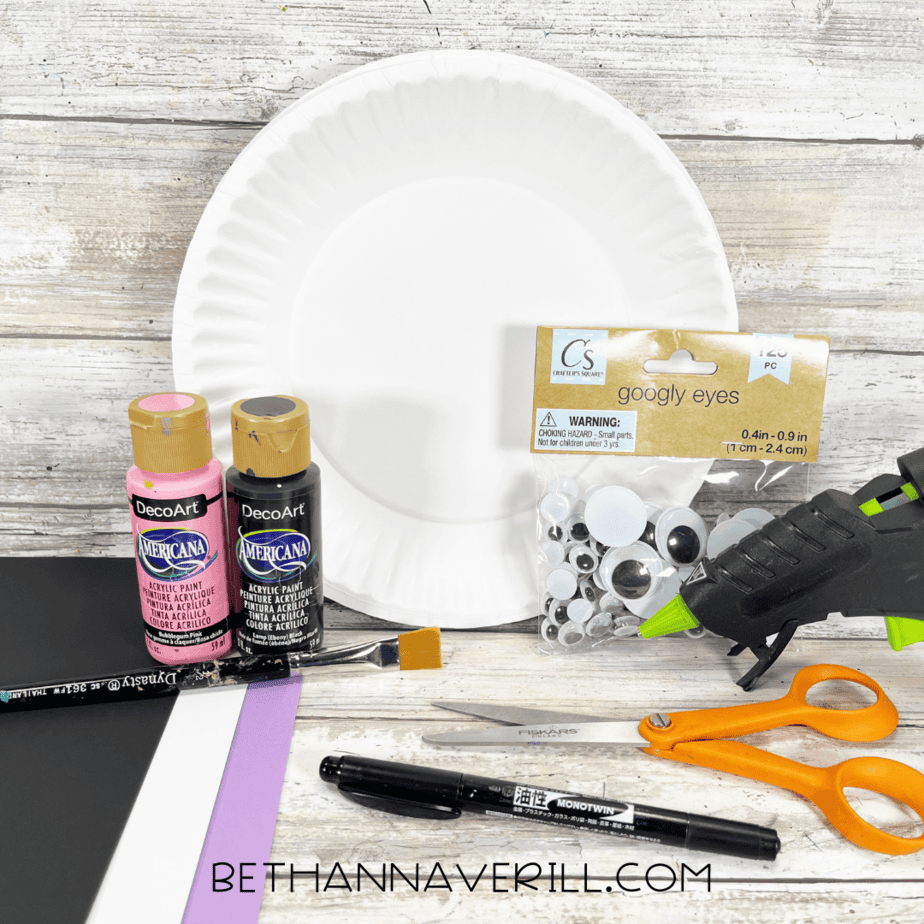 Materials for bat paper plate craft including paper plate, paints, googly eyes, scissors, glue gun, and markers.