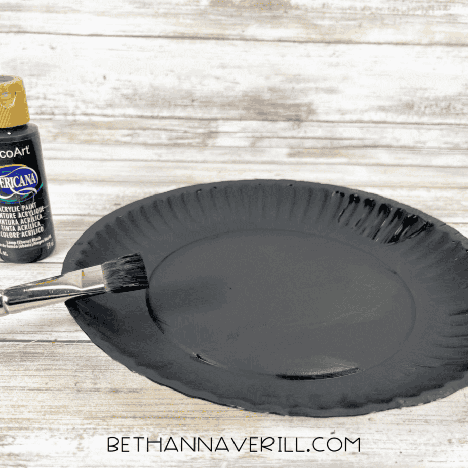 Paper plate painted completely black with paintbrush and bottle of black paint beside it.