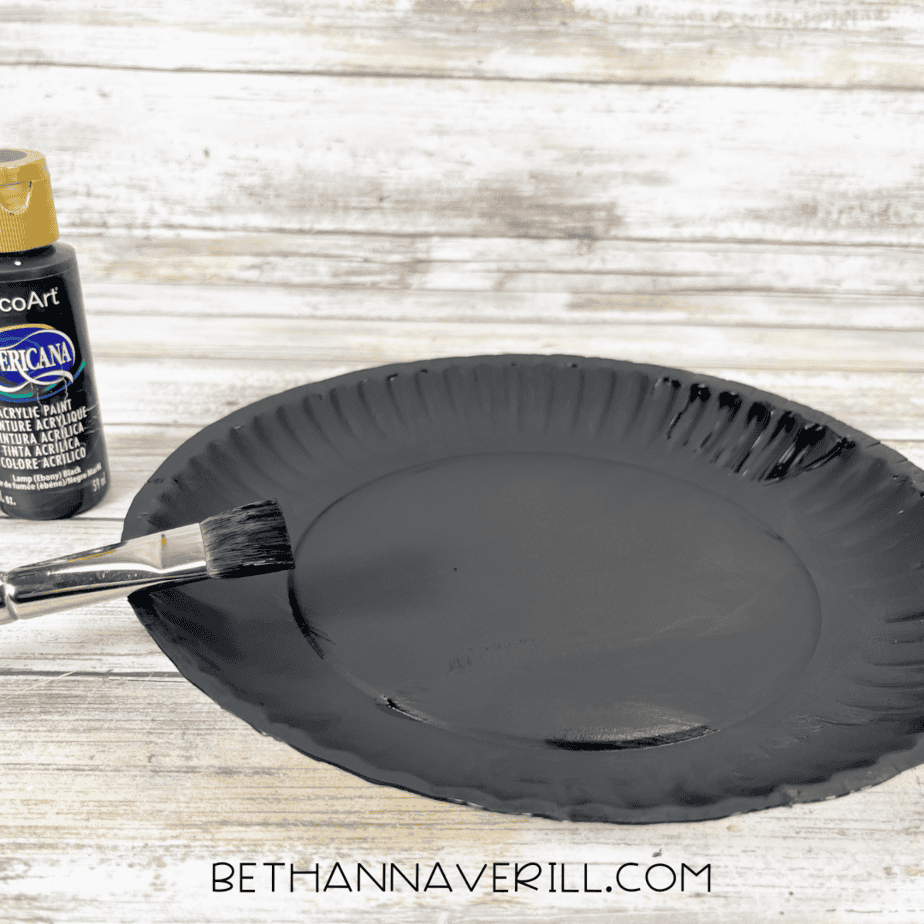 Paper plate painted completely black with paintbrush and bottle of black paint beside it.