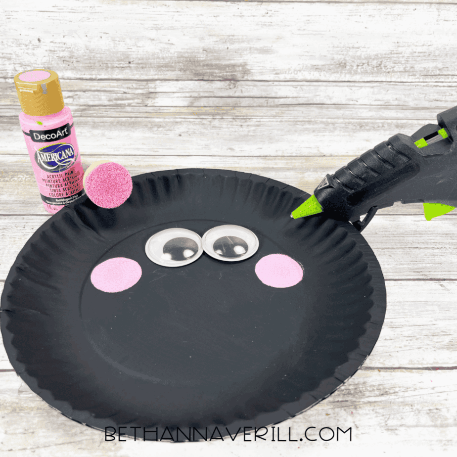 Black painted paper plate with large googly eyes and two pink painted cheeks glued on.