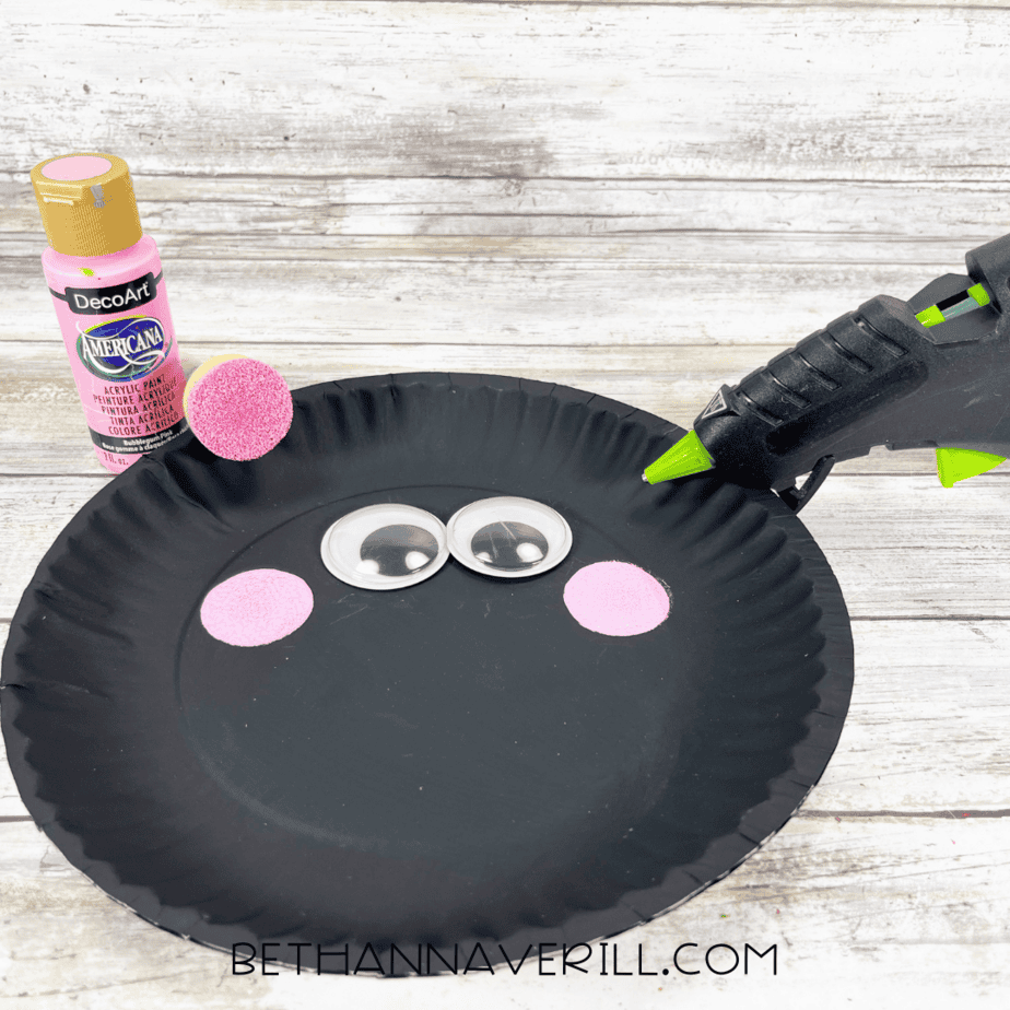 Black painted paper plate with large googly eyes and two pink painted cheeks glued on.