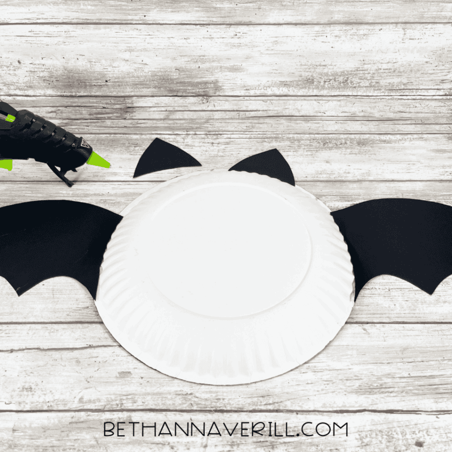Back side of white paper plate showing bat wings and ears being glued on with hot glue gun.