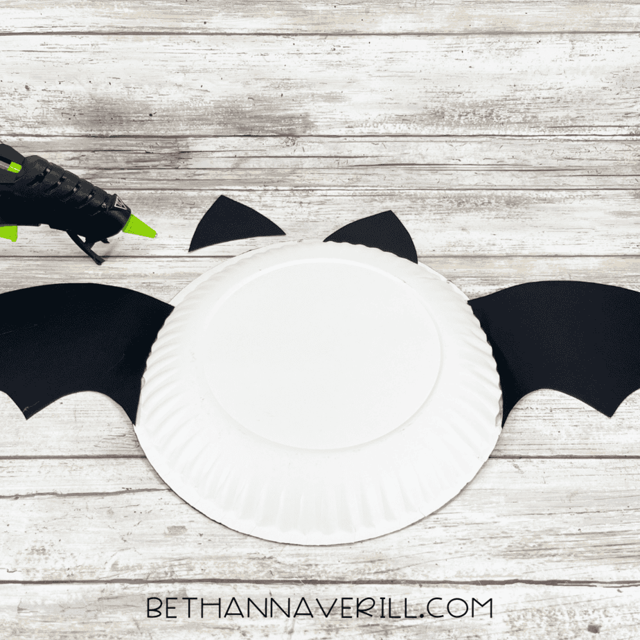 Back side of white paper plate showing bat wings and ears being glued on with hot glue gun.