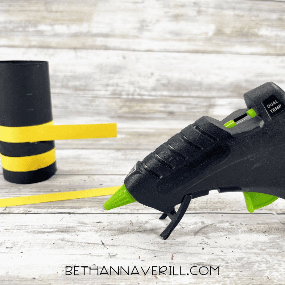 Yellow paper strip being glued around black paper roll with hot glue gun.