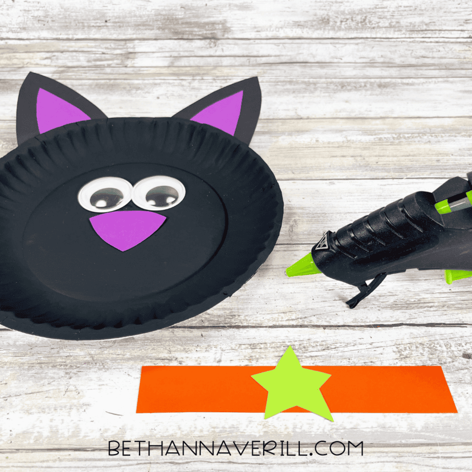 Black painted paper plate with glued-on ears, googly eyes, and purple nose, with orange collar strip nearby.