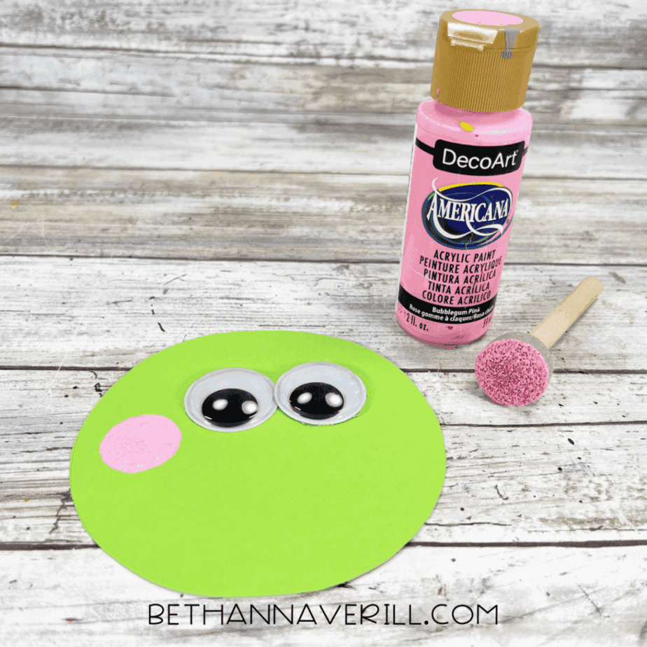 Green paper circle decorated with googly eyes and pink painted cheeks.