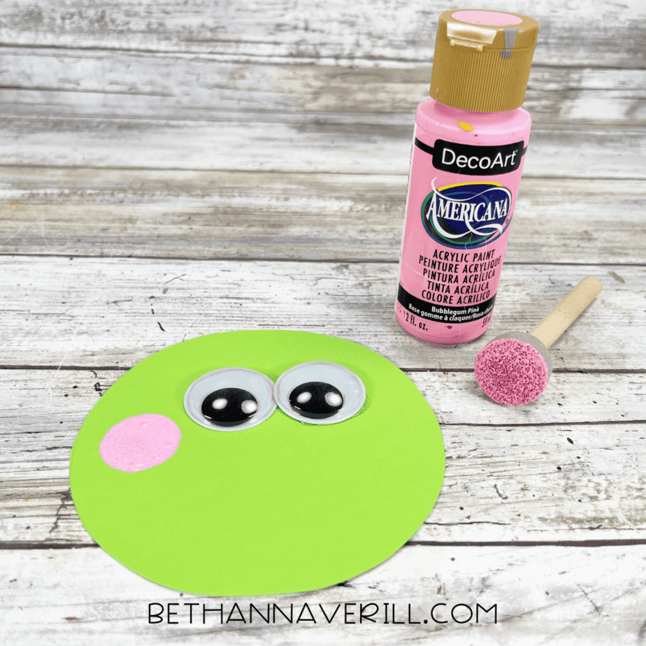 Green paper circle decorated with googly eyes and pink painted cheeks.