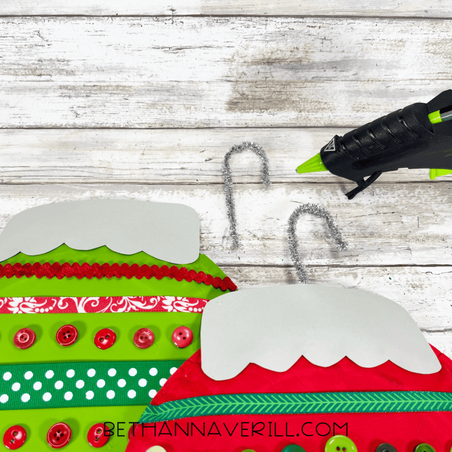 Silver pipe cleaners shaped like hooks being glued onto ornament tops.