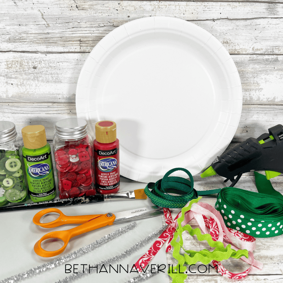 Materials for Christmas ornament paper plate craft including paints, ribbons, scissors, glue gun, and buttons.
