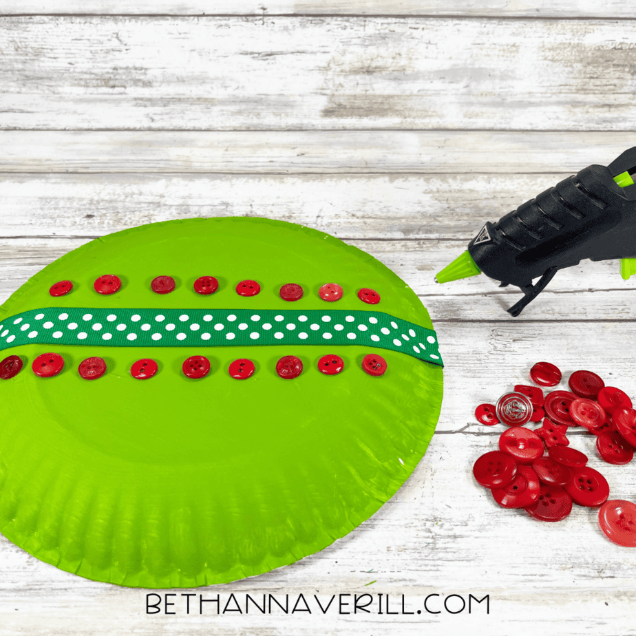 Green painted paper plate decorated with a polka dot ribbon and red buttons being glued on.