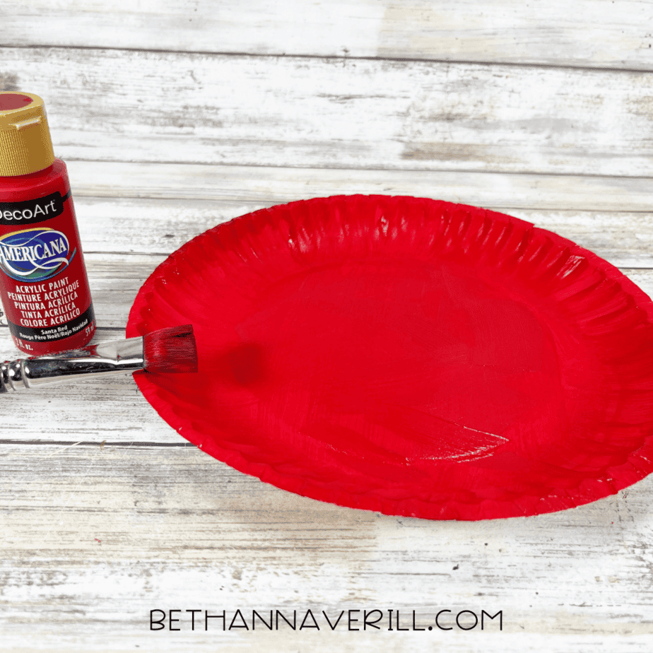 Paper plate painted completely red with paintbrush and red paint bottle nearby.
