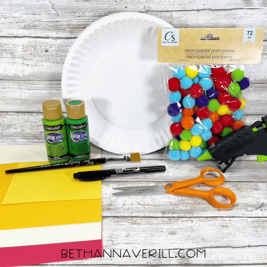 Materials for Christmas wreath craft including paper plate, green paint, pom poms, glue, and scissors.