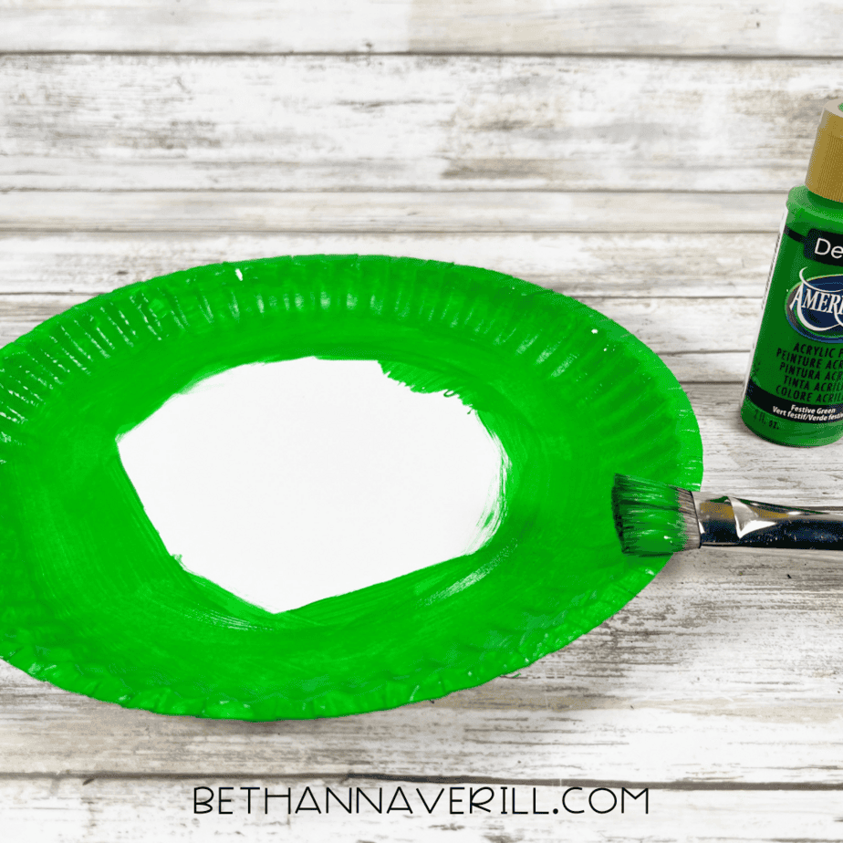Paper plate partially painted green with a brush and paint bottle.