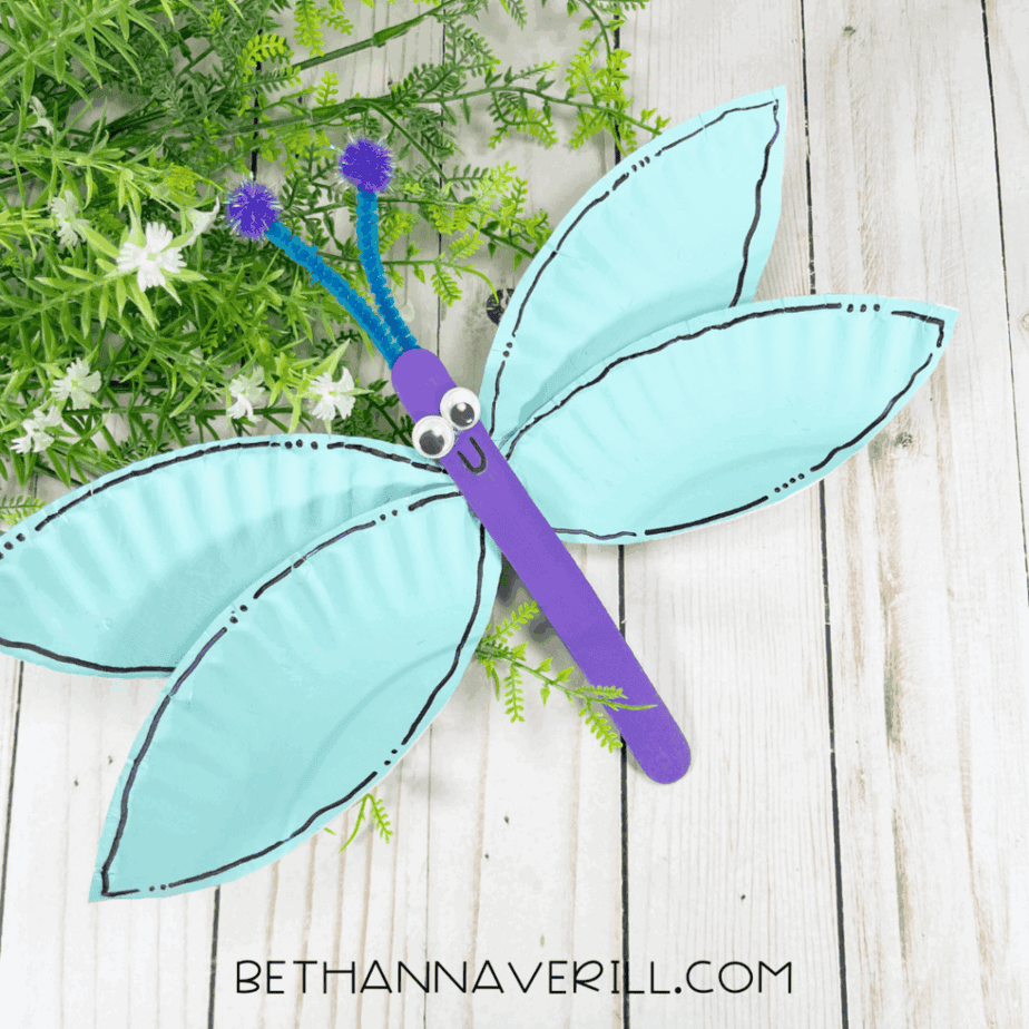 Finished dragonfly paper plate craft with blue wings, purple body, googly eyes, and antennae resting on greenery.