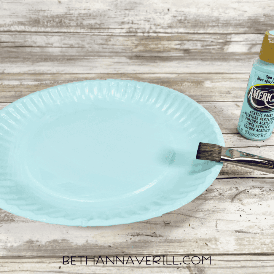 Paper plate painted light blue with a paintbrush and bottle of paint nearby.