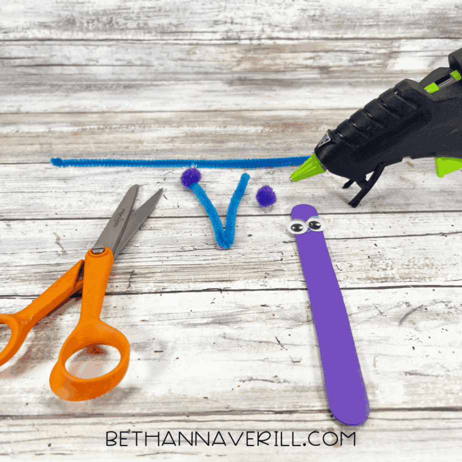 Purple craft stick with googly eyes and blue pipe cleaner antennae next to scissors and glue gun.