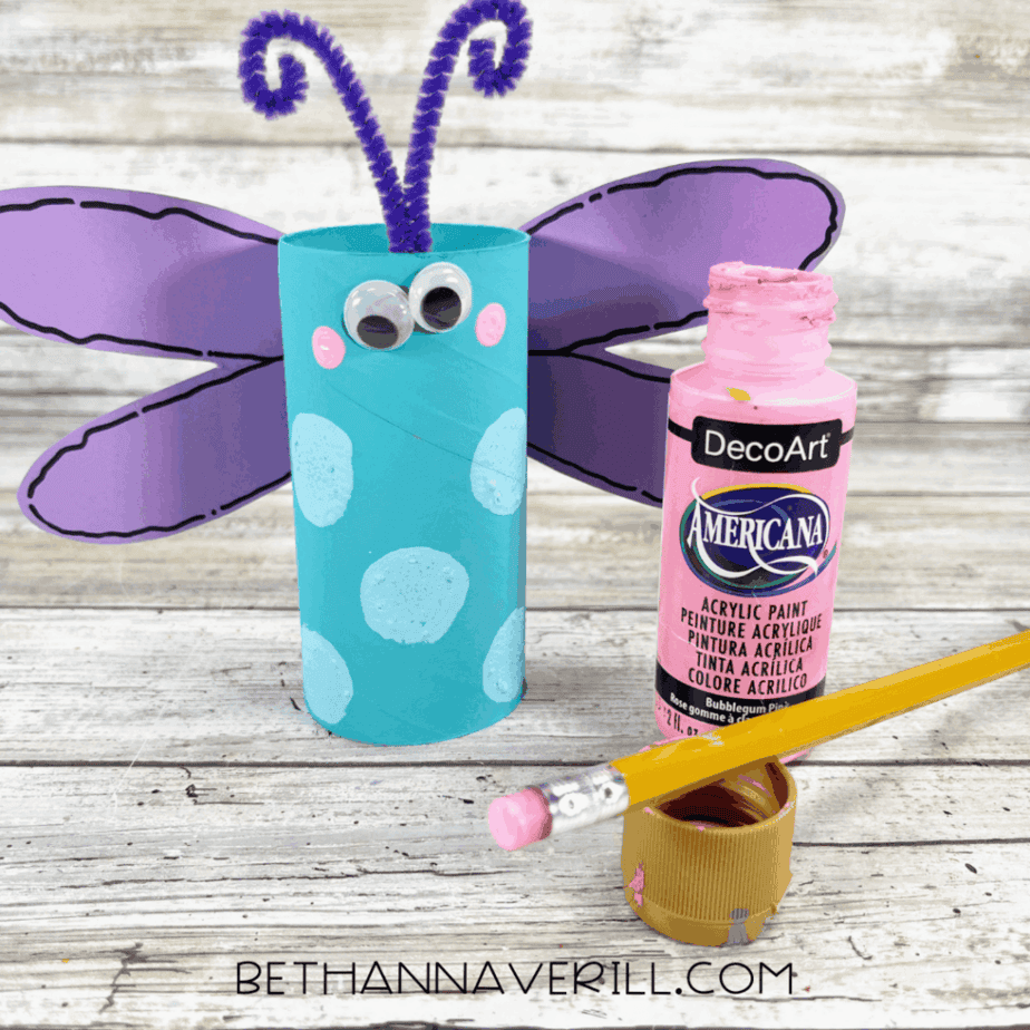 Dragonfly craft with pink painted cheeks using a pencil eraser and paint bottle nearby.