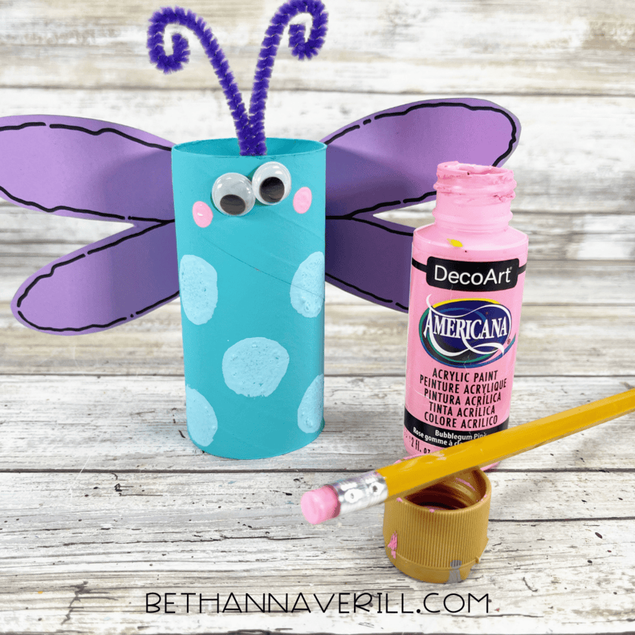 Dragonfly craft with pink painted cheeks using a pencil eraser and paint bottle nearby.