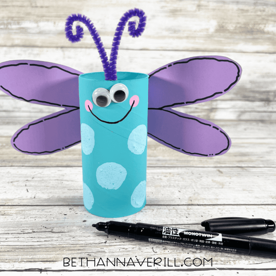 dragonfly paper roll craft with purple wings, googly eyes, rosy cheeks, and a smiling face.