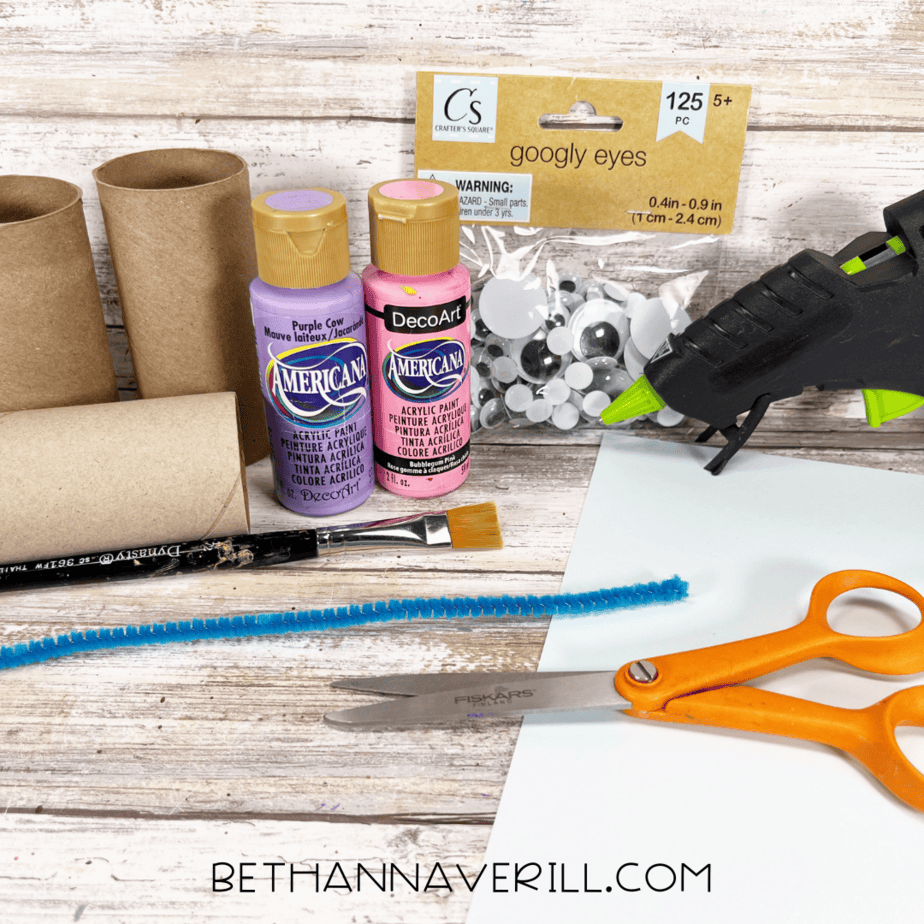Dragonfly paper roll craft supplies including toilet paper rolls, paint, googly eyes, pipe cleaner, scissors, and glue gun.