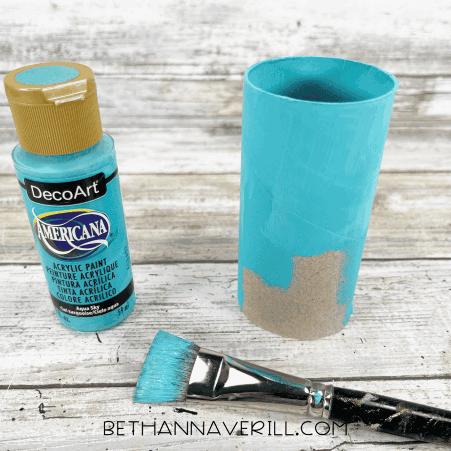 Toilet paper roll painted aqua blue with a paintbrush and paint bottle beside it.