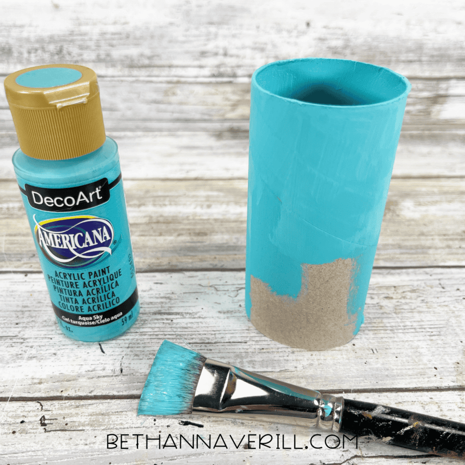 Toilet paper roll painted aqua blue with a paintbrush and paint bottle beside it.