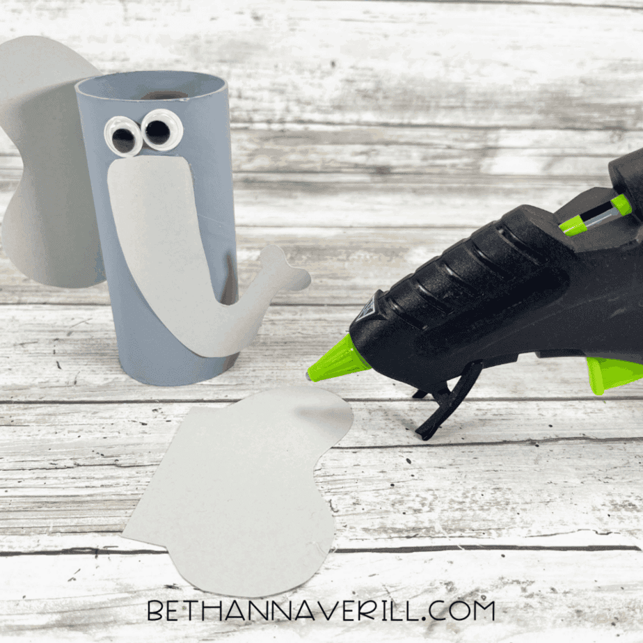 Gray painted toilet paper roll with ears and trunk being glued on, glue gun beside it.