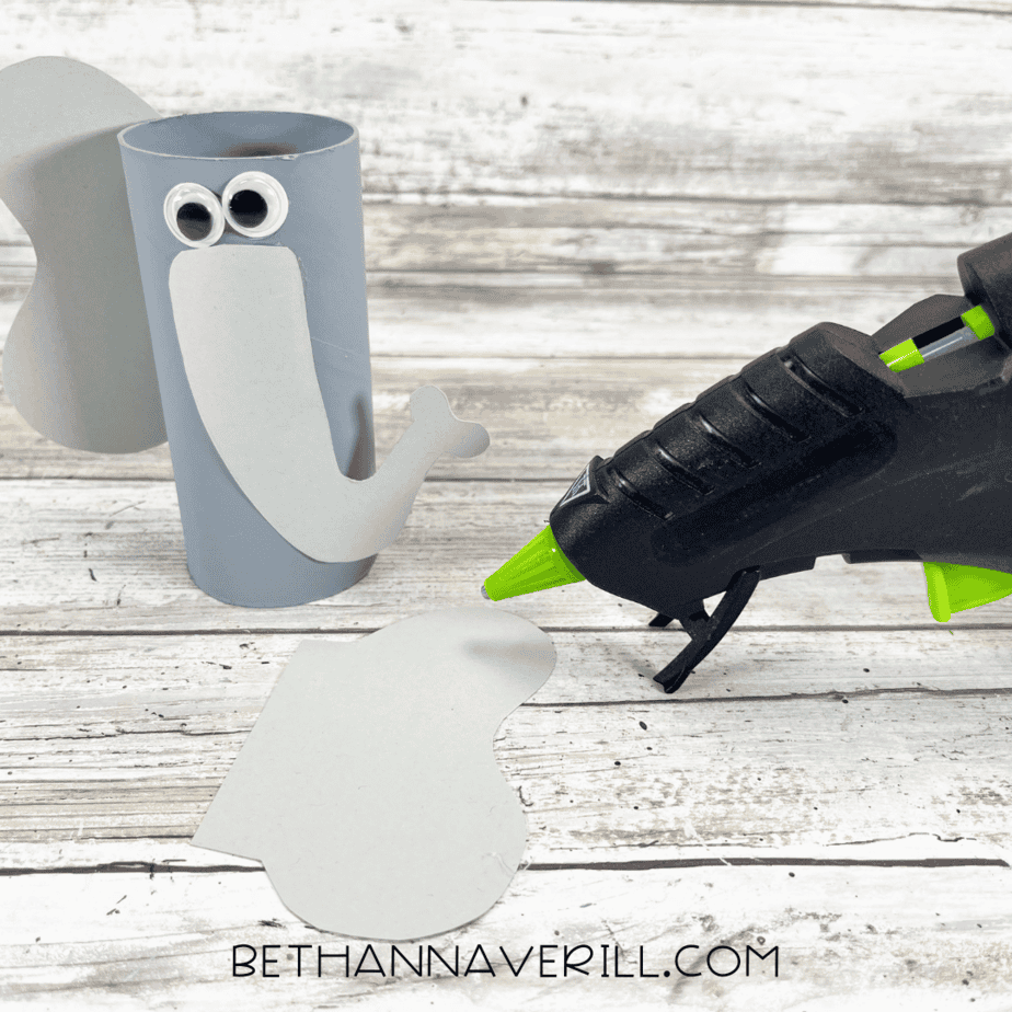Gray painted toilet paper roll with ears and trunk being glued on, glue gun beside it.