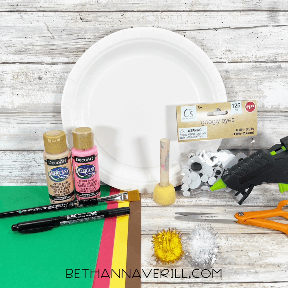 Christmas craft supplies including paper plate, paints, construction paper, pom-poms, googly eyes, and glue gun.