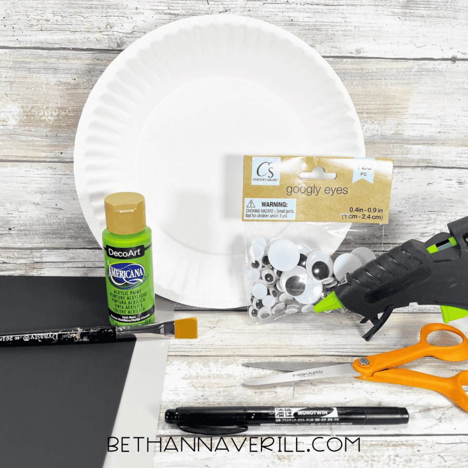 Craft materials including a paper plate, green paint, googly eyes, scissors, glue gun, black and gray paper, and marker.