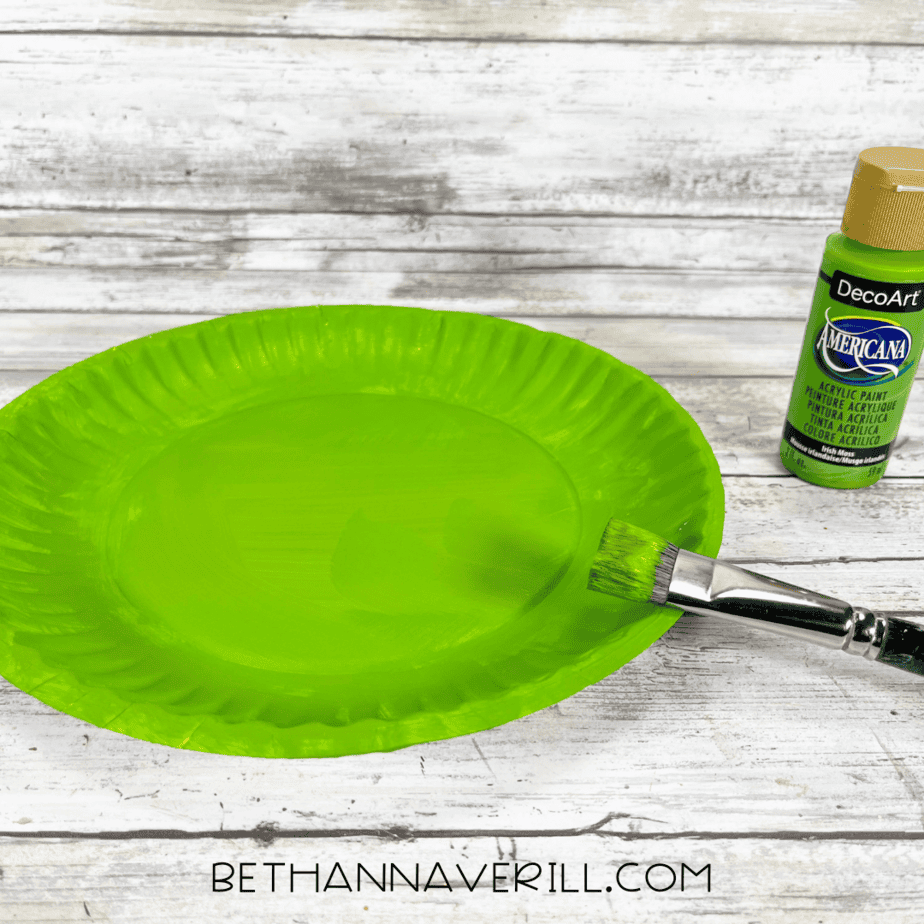 White paper plate painted bright green with a paintbrush and green acrylic paint beside it.