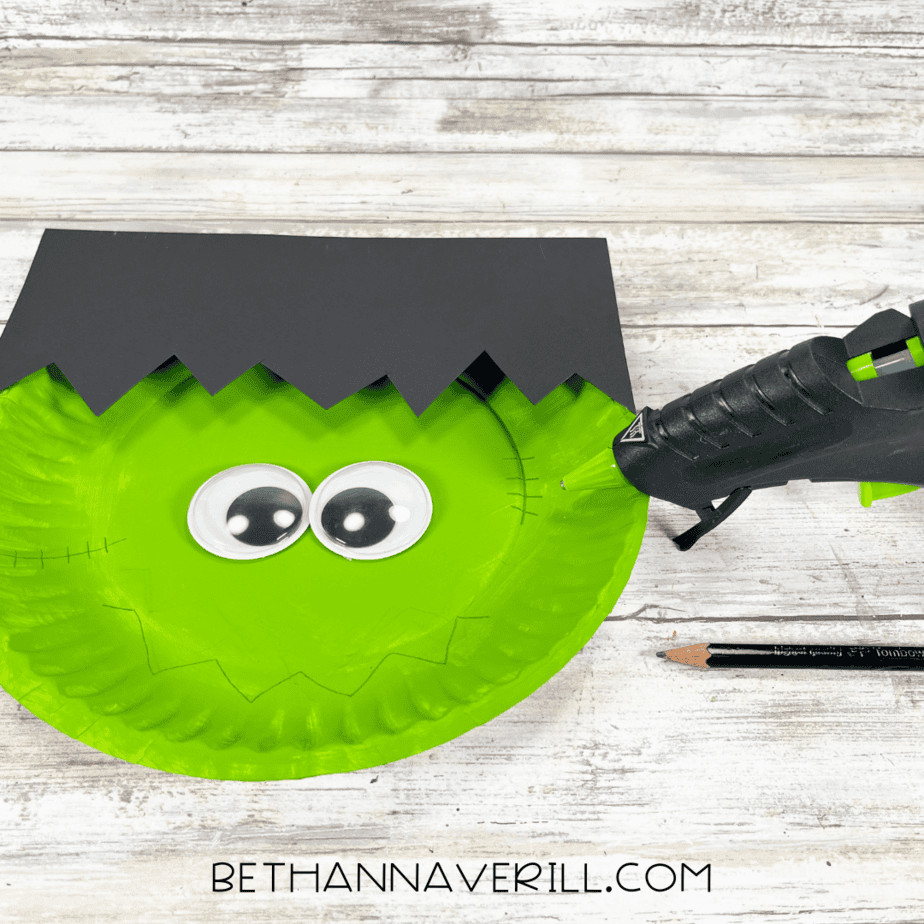 Green painted paper plate with glued-on jagged black hair and googly eyes, glue gun nearby.