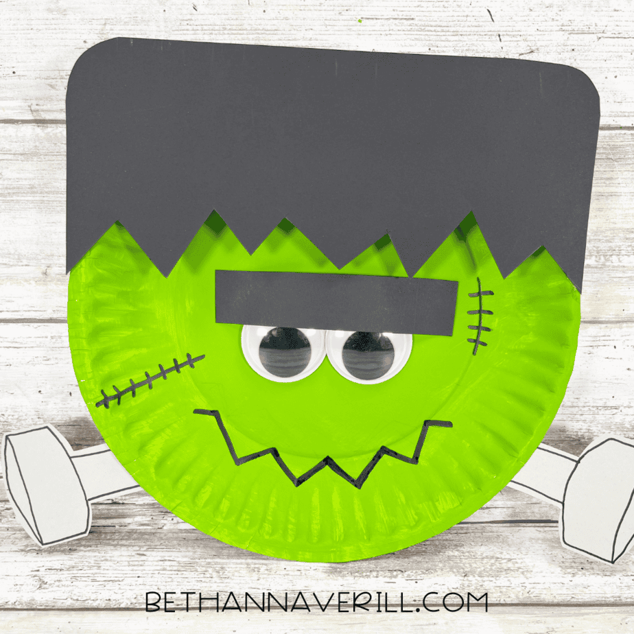 Finished Frankenstein paper plate craft with green face, black hair, stitches, googly eyes, and gray bolts.