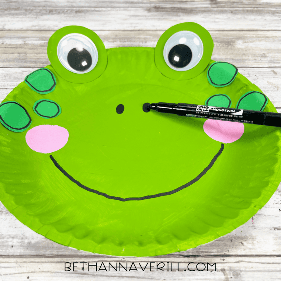 A black marker drawing nostrils and a large smiling mouth on the frog’s face.