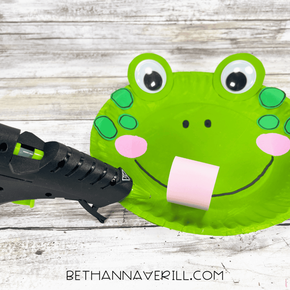 A glue gun attaching the curled pink tongue to the bottom of the frog’s smiling mouth.
