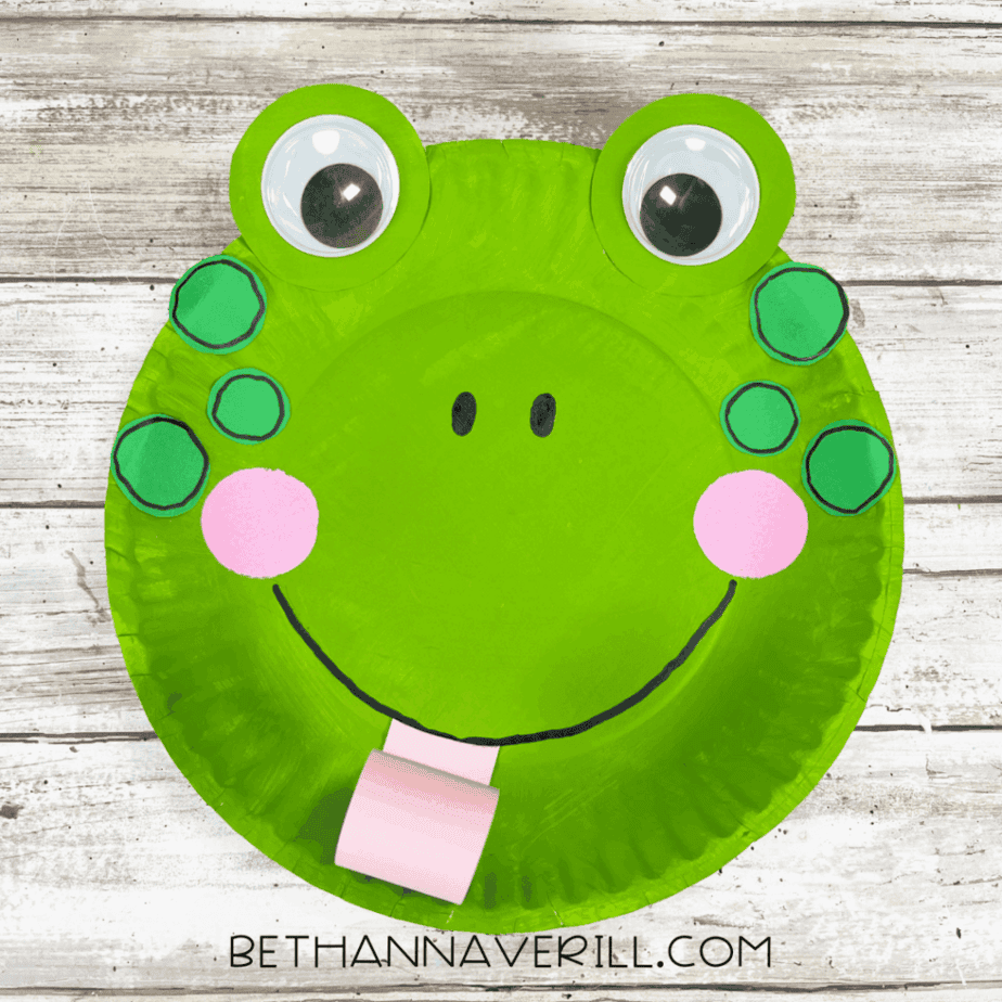 A finished frog paper plate craft with big googly eyes, green spots, rosy cheeks, a smile, and a curly pink tongue.
